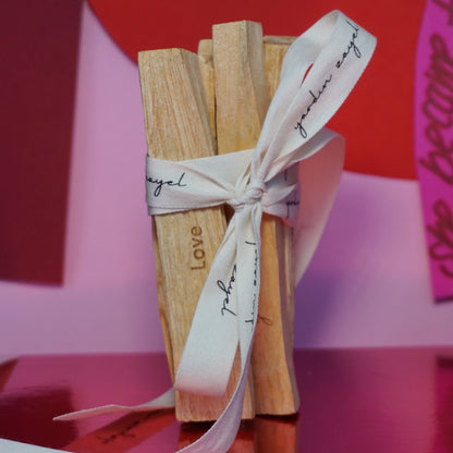 Palo Santo Bundle aus 5 Sticks mit dem Mantra Love sind auf einer pinken fläche zu sehen. Das Set ist zusammengebunden mit einer beigen Schnur, auf welchem yardin zayel in Schwarz zu sehen ist